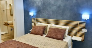 A bedroom with a wooden headboard, two bedside lamps, a bed with a striped bedspread, and a bathroom with a sink and mirror. - La Piccola Perla Rooms (Sant'Agnello)