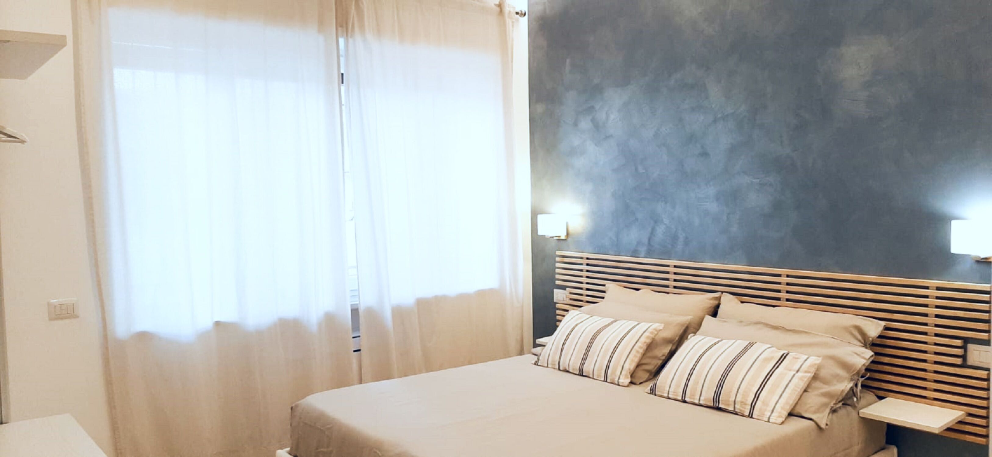 Un dormitorio con cama, cortinas, una mesita de noche y una lámpara fijada a la pared.