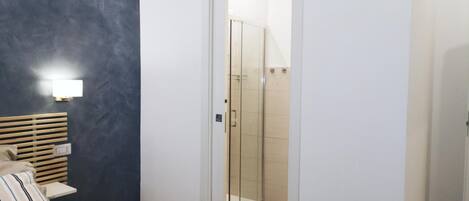 Ein modernes Schlafzimmer mit einem Bett, Nachttisch und einem kleinen Tisch daneben. Eine Glastür führt zur Badezimmerecke mit Dusche und Toilette.