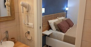 A hotel room with a wooden headboard, a bedside table, a white bed with beige and brown pillows, a mirror with a gold frame, and a hairdryer mounted on the wall. - La Piccola Perla Rooms (Sant'Agnello)