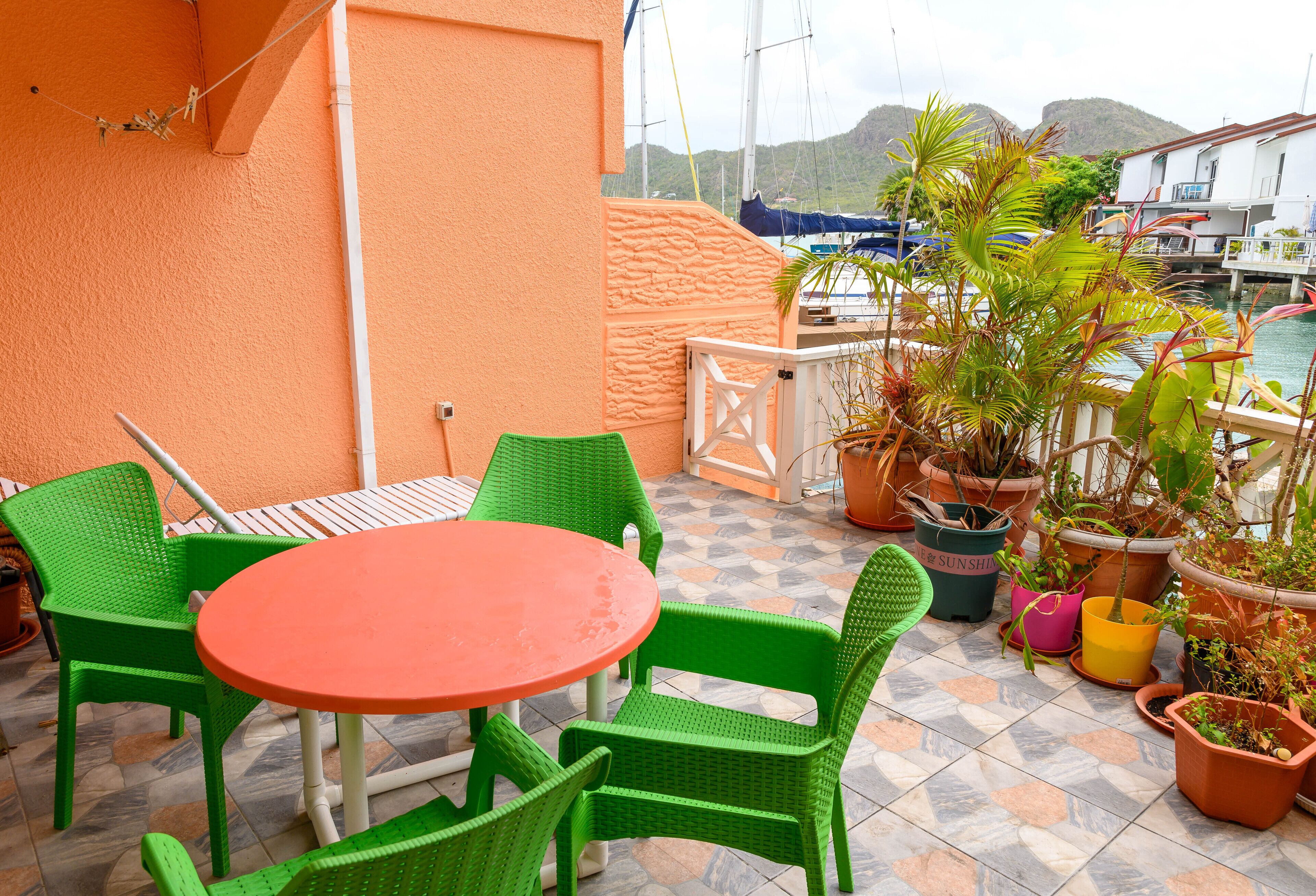 Un patio con una mesa redonda y sillas verdes, rodeado de plantas en macetas y vistas a barcos y colinas al fondo.