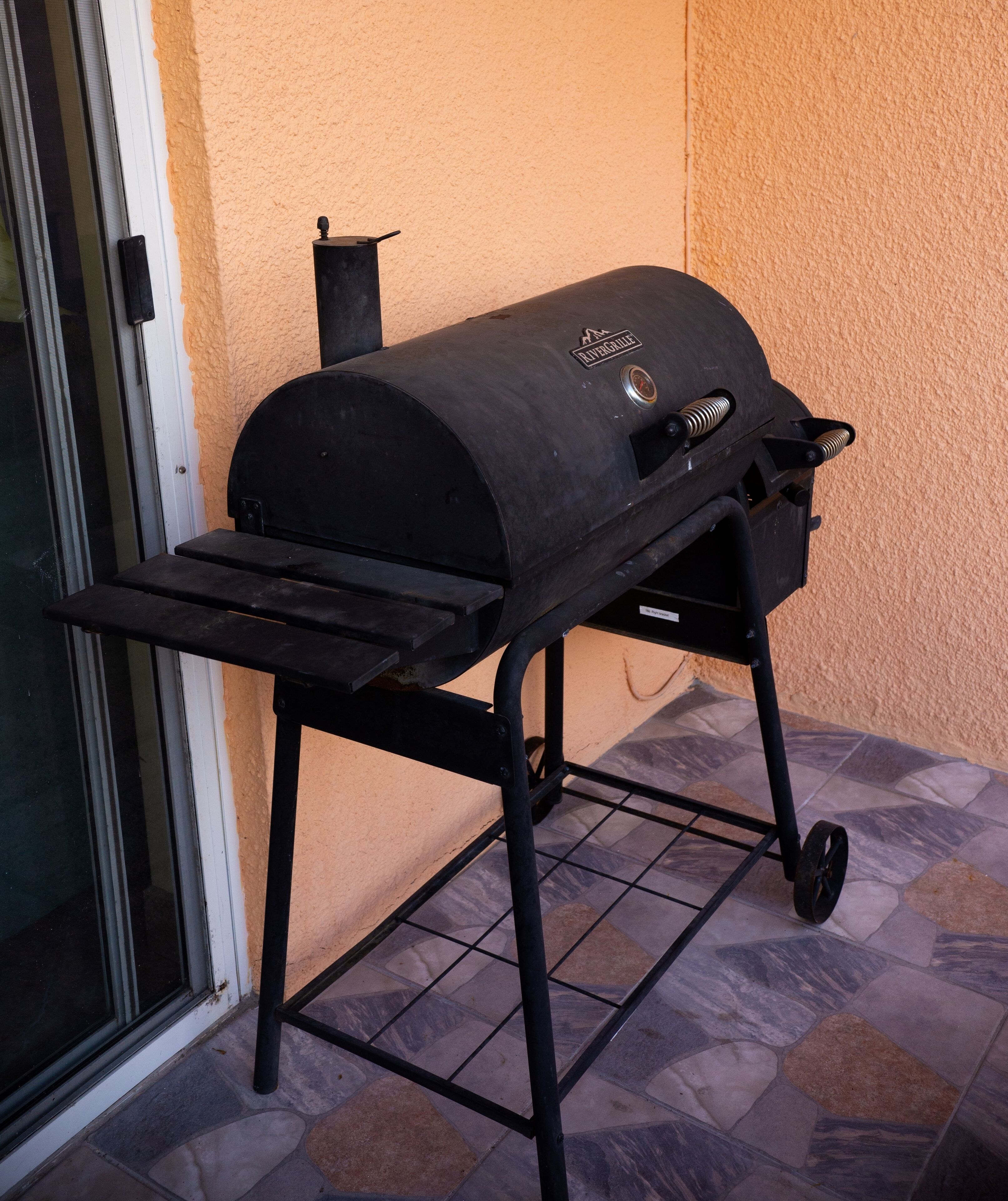 Ein schwarzer Grill mit Rollen steht auf einer gefliesten Terrasse neben einer Glastür.