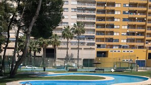 Une piscine au design incurvé, entourée d’une terrasse en béton et d’une pelouse. Au fond, on aperçoit un immeuble à plusieurs étages doté de balcons.