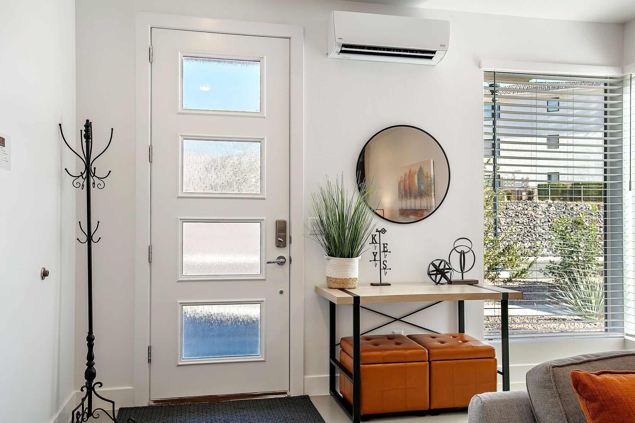 Un salón moderno con una puerta blanca, un perchero de metal negro, un espejo redondo, una mesita con una planta y vistas a un jardín a través de amplios ventanales.