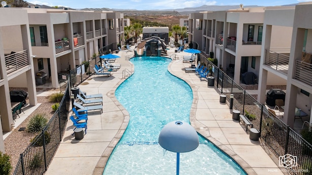 A swimming pool surrounded by multi-story buildings with balconies, lounge chairs, and a clear sky.