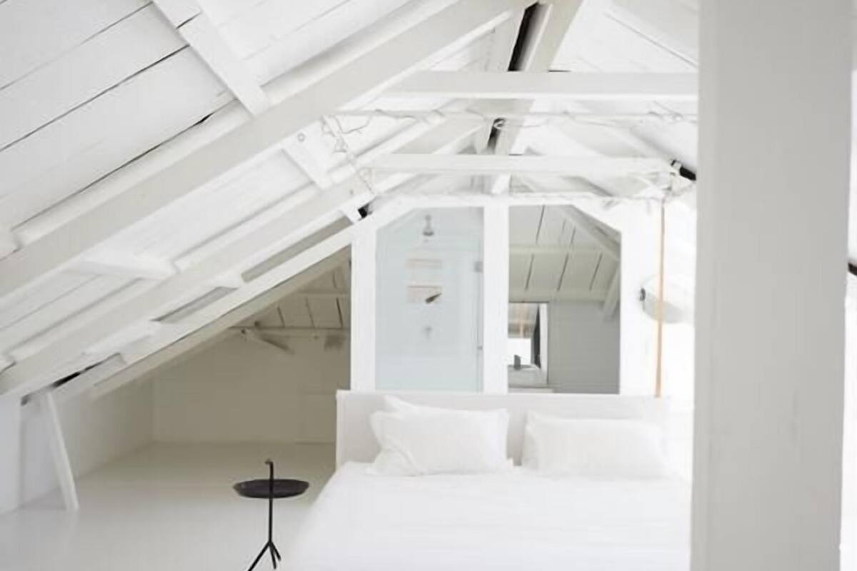 Une chambre à coucher moderne, aux tons blancs, avec un plafond en pente, un lit recouvert de linge de lit blanc et un tapis rayé noir et blanc.