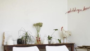 Uma sala de estar minimalista com duas poltronas brancas, uma mesa de centro de madeira e um banquinho branco pequeno. Também se vê um galo decorativo e uma parede com uma mensagem escrita à mão.