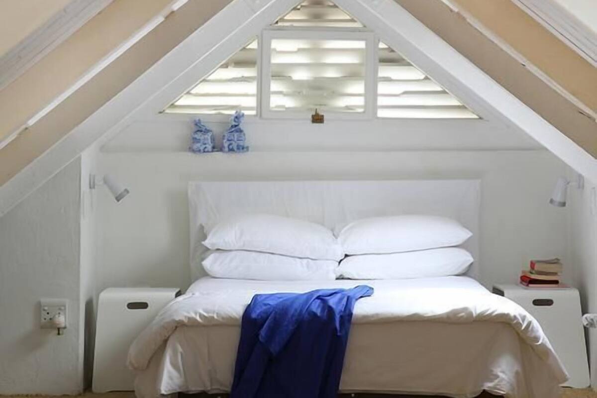 Une chambre avec un plafond en pente, un lit avec une literie blanche, une couverture bleue et une paire de chaussures sur le sol.