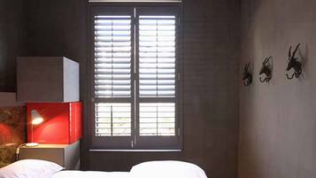 Un dormitorio con cama, mesita de noche, lámpara, ventana con persianas y una obra de arte colgada en la pared.
