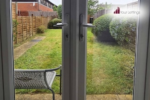 A view through a sliding glass door into a backyard with a lawn, a wooden fence, and a metal chair.