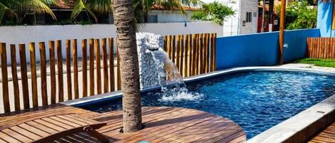 Zwembadgedeelte met houten terras, lounge stoelen en een kleine waterval.