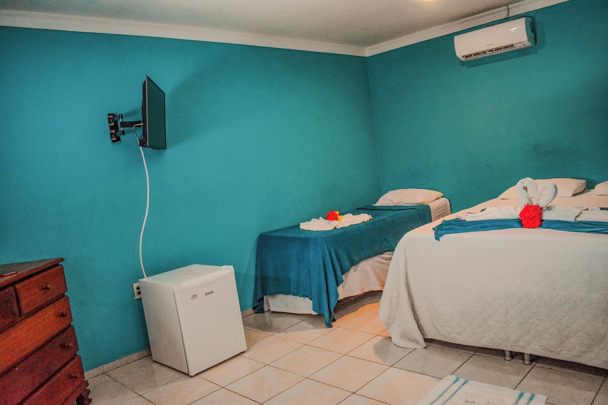 Ein Zimmer mit türkisfarbenen Wänden, einem Bett mit blauer und weißer Bettdecke, einem kleinen Nachttisch mit Lampe, einer weißen Klimaanlage und einem hölzernen Schminktisch.