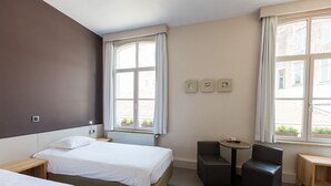A hotel room with two beds, a small table, a chair, and large windows.
