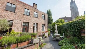 A brick building with a garden patio, outdoor seating, and a view of a tall church tower.