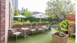 Een binnentuin met een bakstenen muur, groen kunstgras, potplanten en zitplaatsen met parasols.