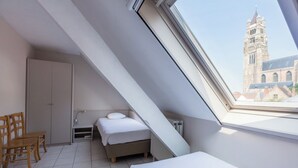 Une chambre avec un plafond en pente, un lit, une chaise, une armoire et une vue sur une tour par la fenêtre.