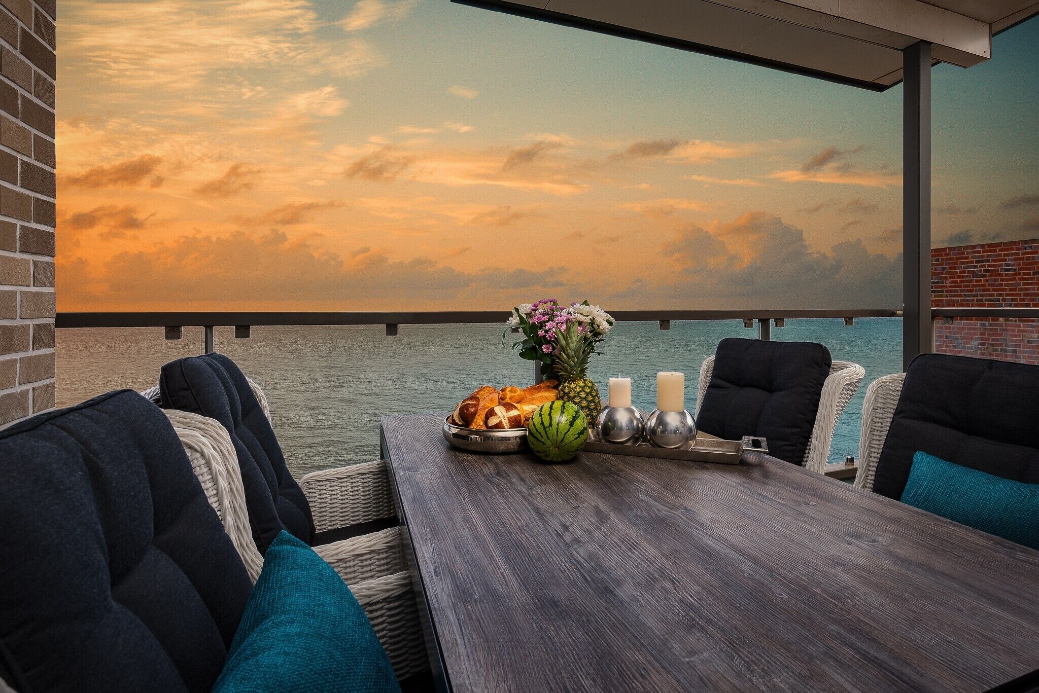 Un balcon agrémenté de meubles en osier, une table en bois avec un panier de fruits, des bougies et une vue sur le coucher de soleil sur l’eau.