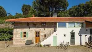 A stone house with a red-tiled roof, a wooden door, and a staircase leading to a second floor. There are bicycles parked outside and a bench in the front yard.