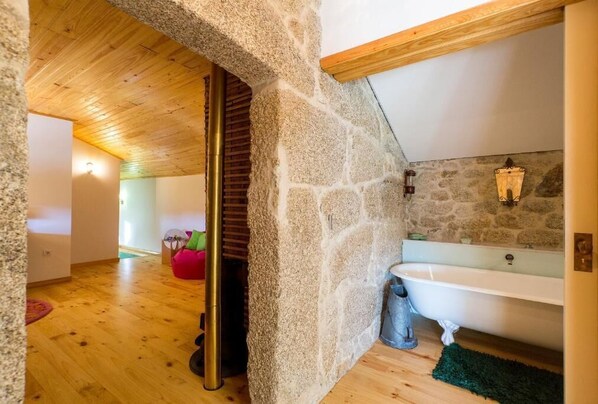 A stone-walled bathroom with a wooden ceiling, a bathtub, and a stone wall with a light fixture.