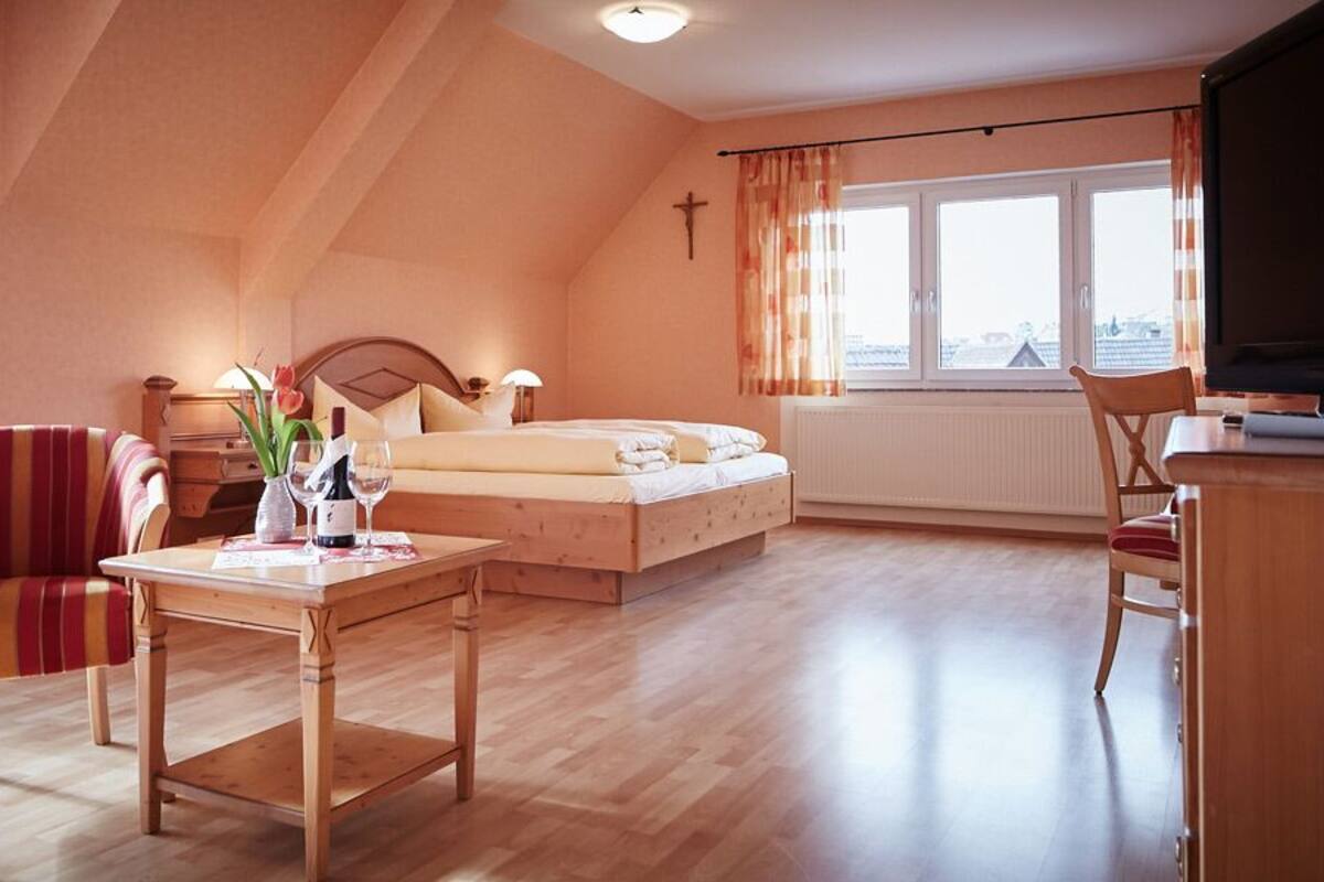 A cozy bedroom with a bed, a wooden table with a vase and a glass, a TV, and a window with curtains.
