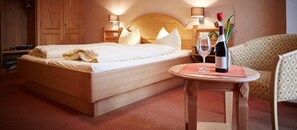A hotel room with a wooden bed, a small round table with a wine bottle and glass, and a chair with a patterned cushion. - Metzgereigasthof Rebstock (Ettenheim)