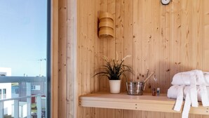 A wooden sauna room with a bench, a shelf with towels, a plant, and a clock on the wall. - Vacation home "Nis Randers" with floor-to-ceiling windows, sauna & view of the Schlei (Olpenitz)