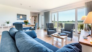 A modern living room with a sofa, armchairs, a dining area, and large windows offering a view. - Vacation home "Nis Randers" with floor-to-ceiling windows, sauna & view of the Schlei (Olpenitz)