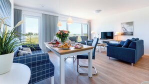A modern living room with a sofa, dining table, and TV. - Vacation home "Nis Randers" with floor-to-ceiling windows, sauna & view of the Schlei (Olpenitz)