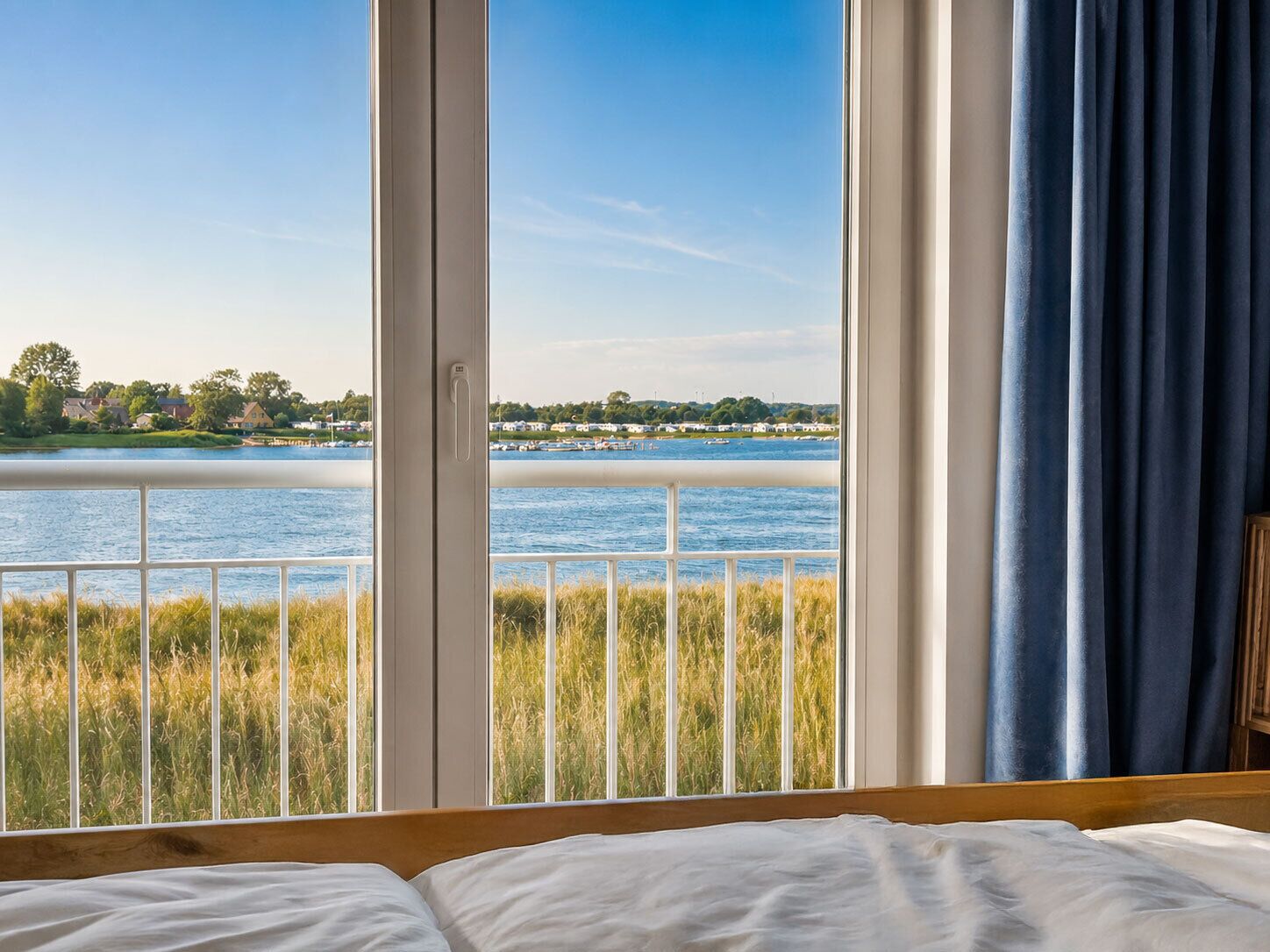 Une chambre avec une grande fenêtre donnant sur un lac, un lit et une vue sur les maisons situées de l’autre côté de l’eau.