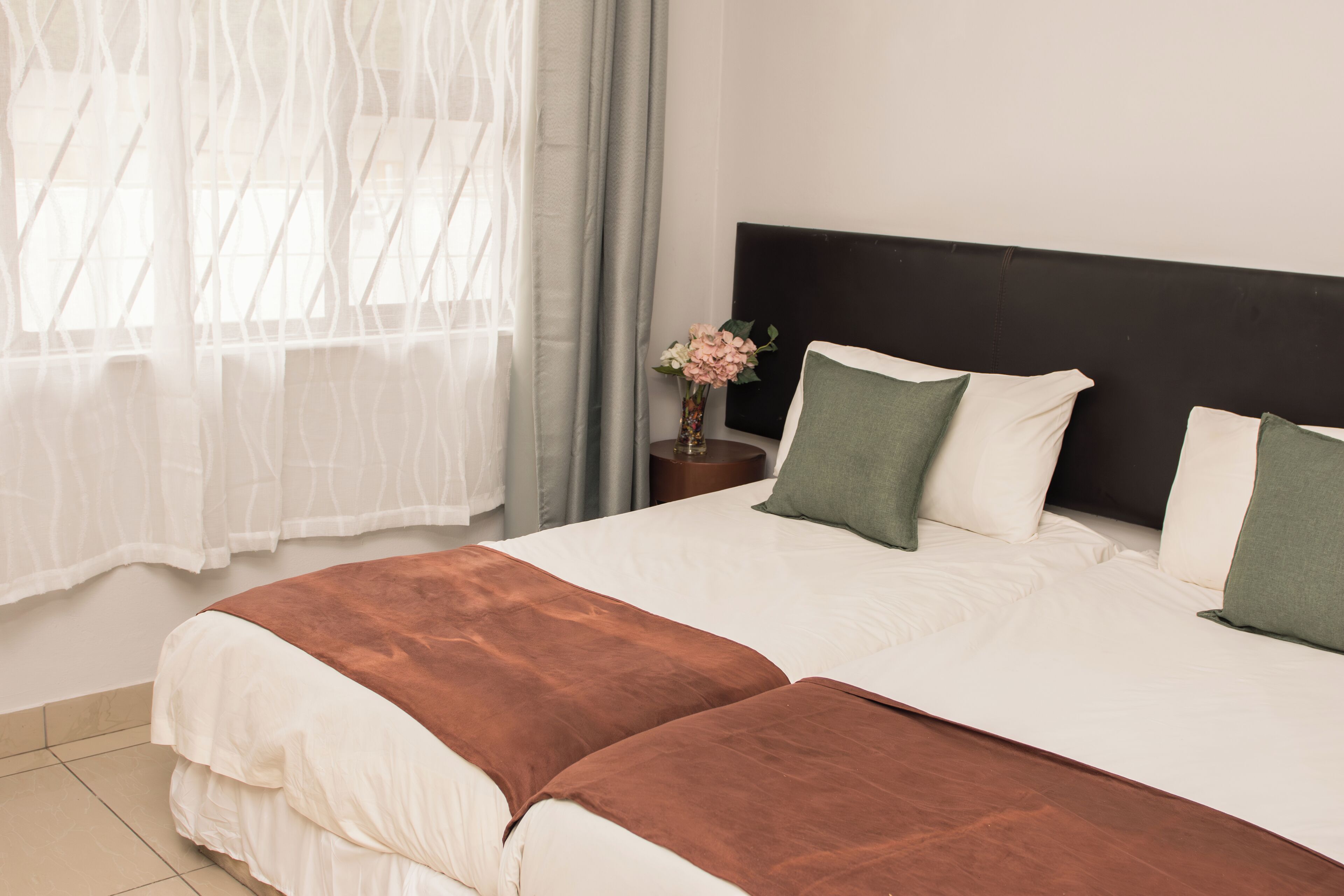 A neatly made bed with white and brown bedding, a dark headboard, and a vase with flowers on a bedside table.