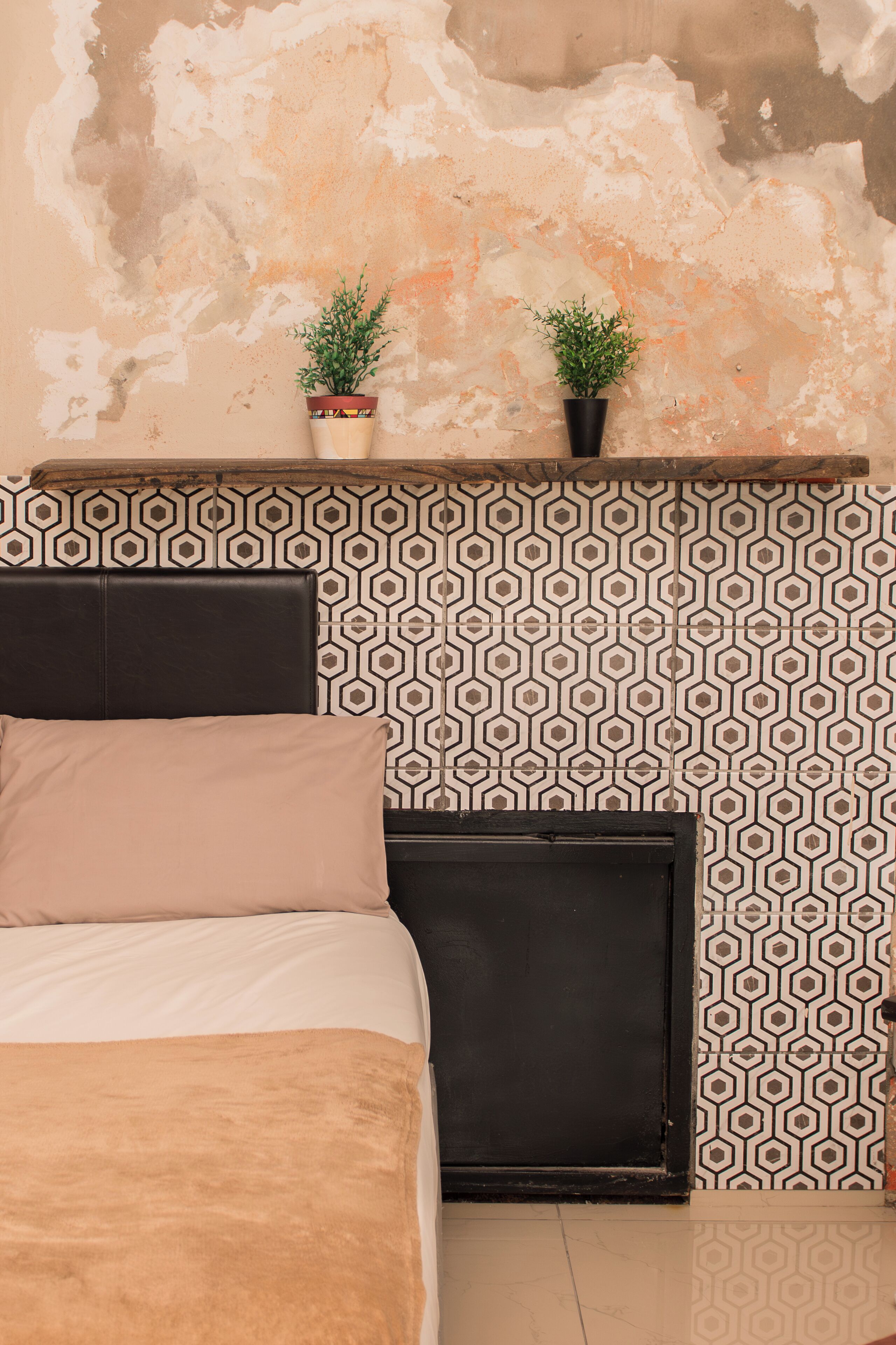 A bedroom with a patterned wall, two potted plants, a bed with a headboard, and a black wardrobe.