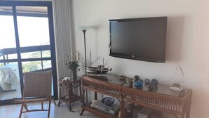 A living room with a flat-screen TV, wicker furniture, a glass coffee table, and a view through a glass door. - Beautiful fit, standing on the sand with spectacular sea views. Spotless (Bertioga)