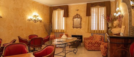 A dining area with a round table, red upholstered chairs, and a sofa. There are wall sconces, a fireplace, and a mirror on the wall.