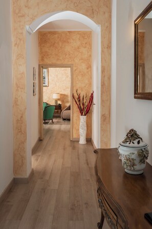 A hallway with a wooden floor, a white vase with dried plants, a green chair, and a wooden cabinet.
