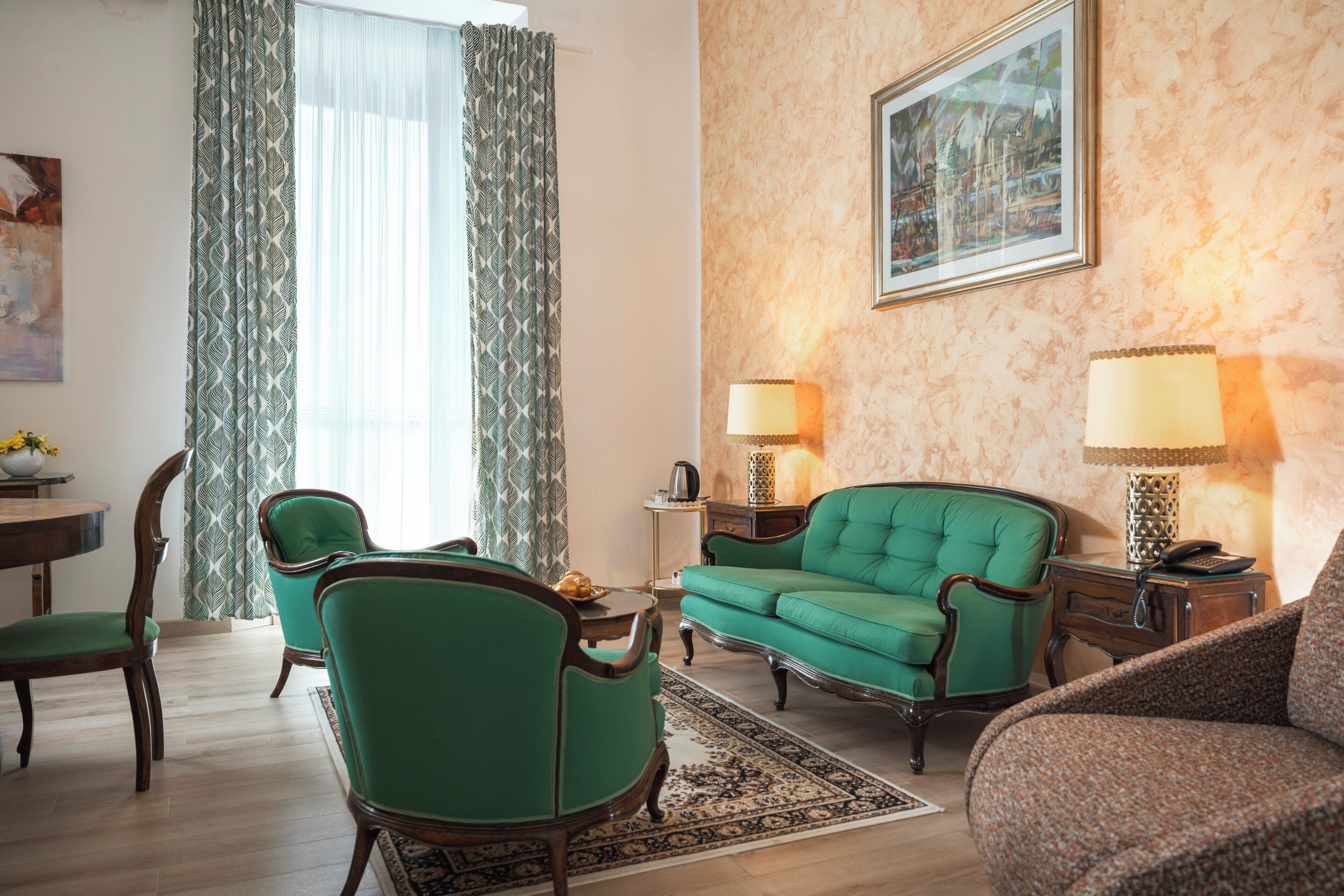 Ein Wohnzimmer mit grünen Polstersesseln, einem Sofa, einem Couchtisch und einem Teppich. An der Wand hängt ein Gemälde und auf einem Beistelltisch steht eine Lampe.