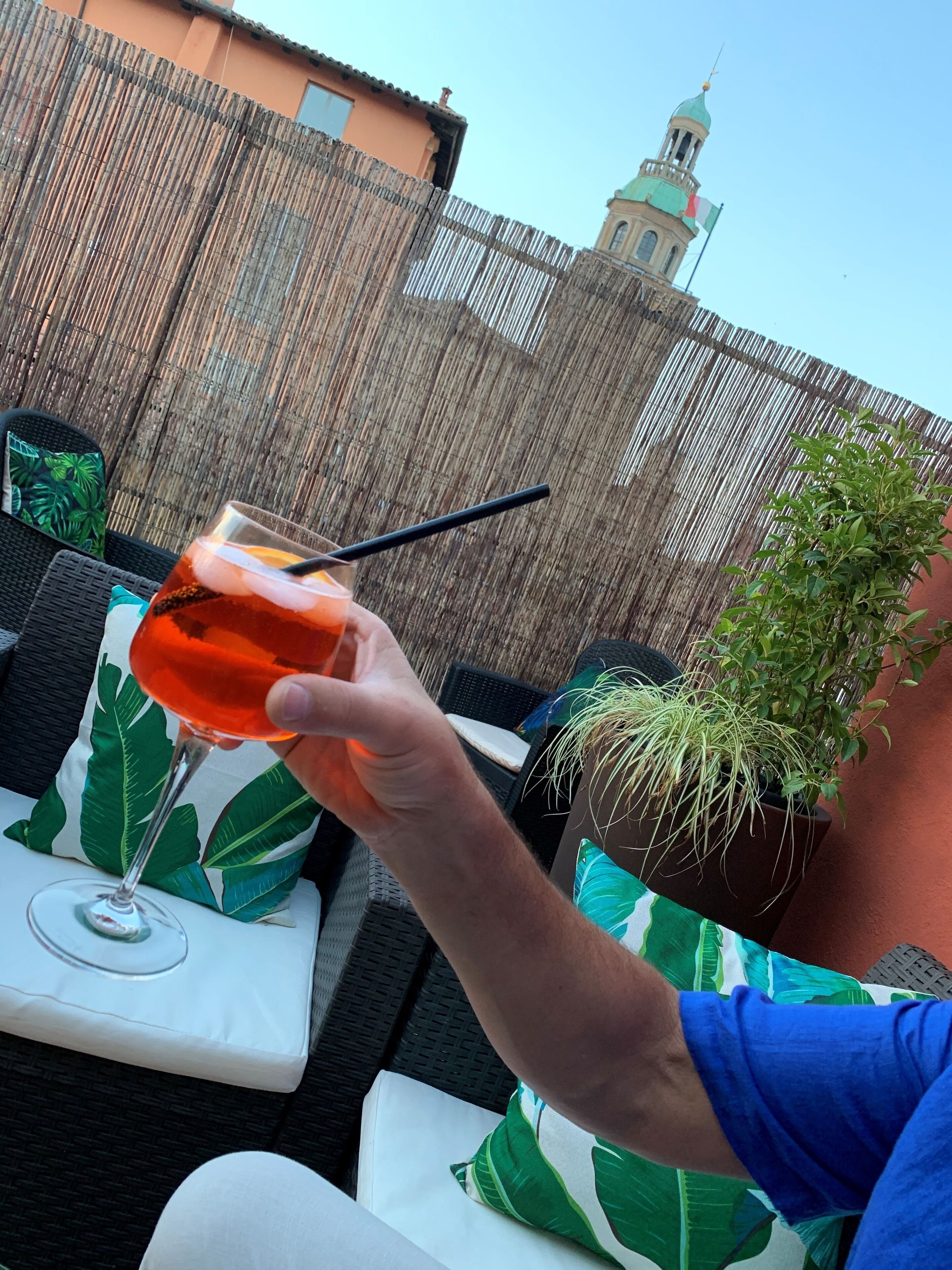 Une personne tenant un verre à cocktail avec une paille, assise à une table agrémentée d’un coussin orné de motifs tropicaux.