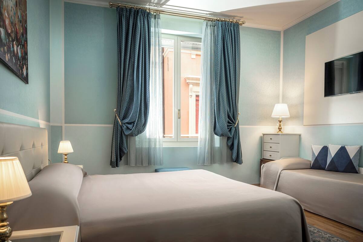 Habitación de hotel con dos camas, una ventana con cortinas, un televisor en la pared y una lámpara en la mesita de noche.