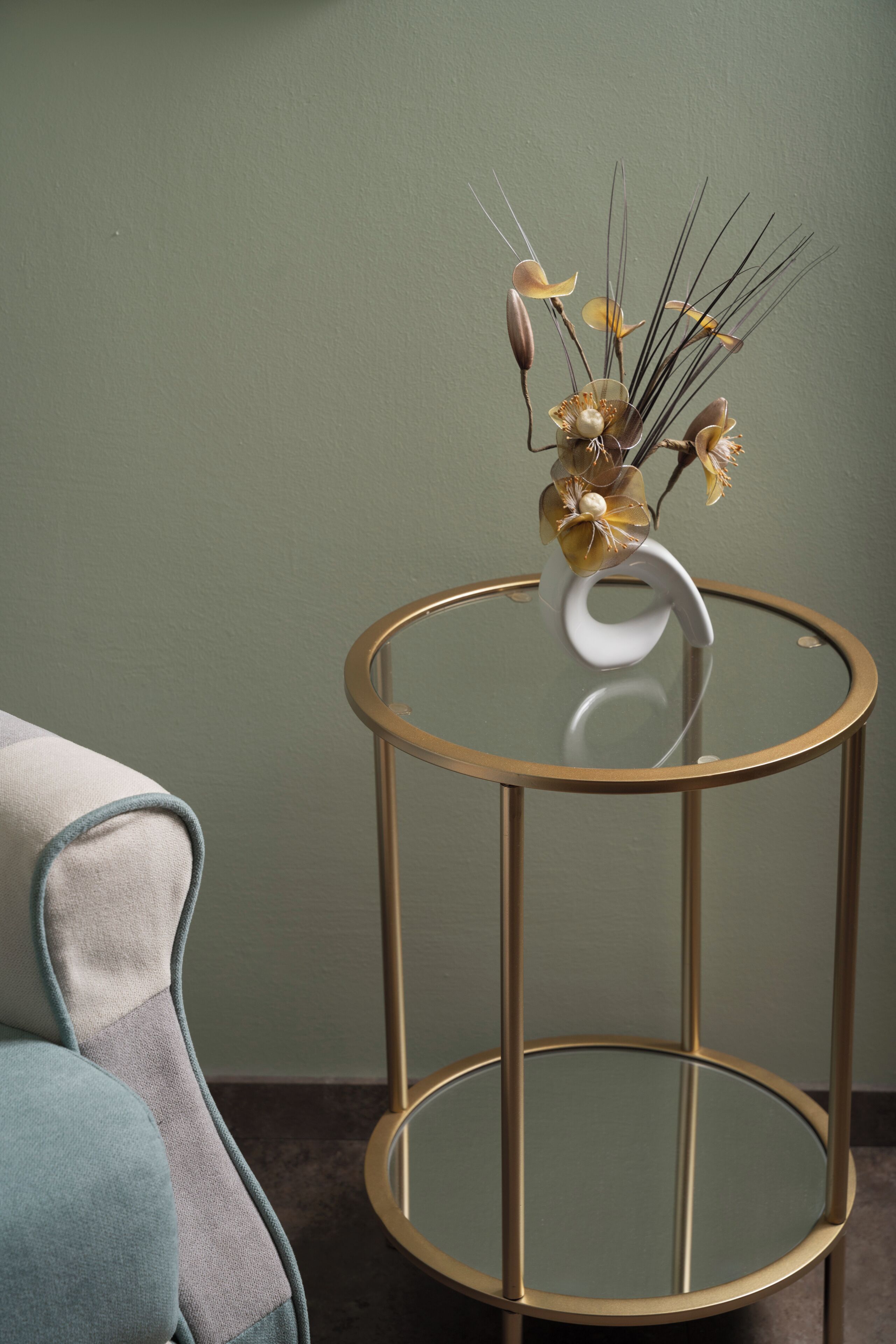 A round glass table with a gold frame, a vase with dried flowers, and a blue chair.