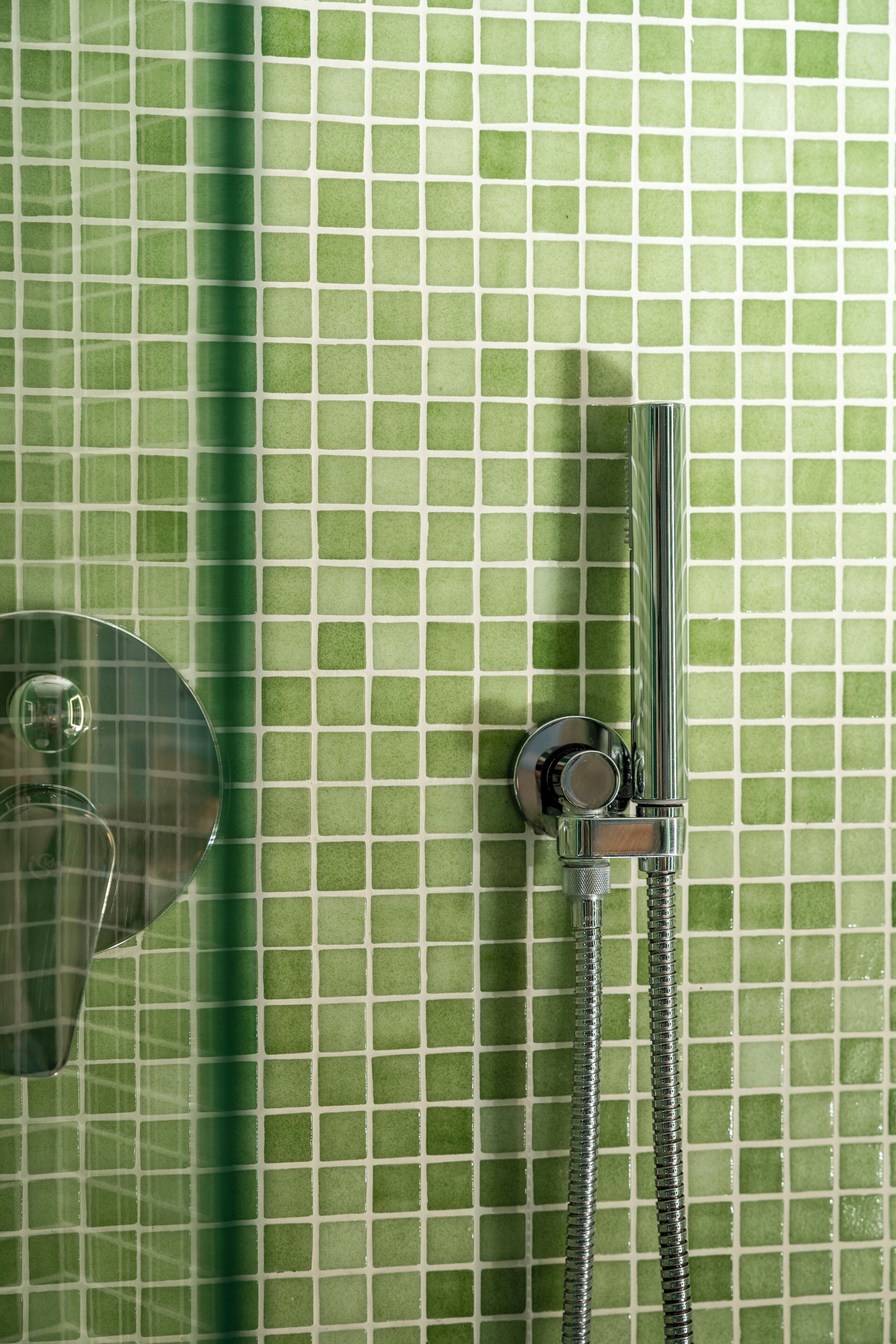 Ducha con grifo y manguera de cromo, frente a una pared de azulejos verdes.