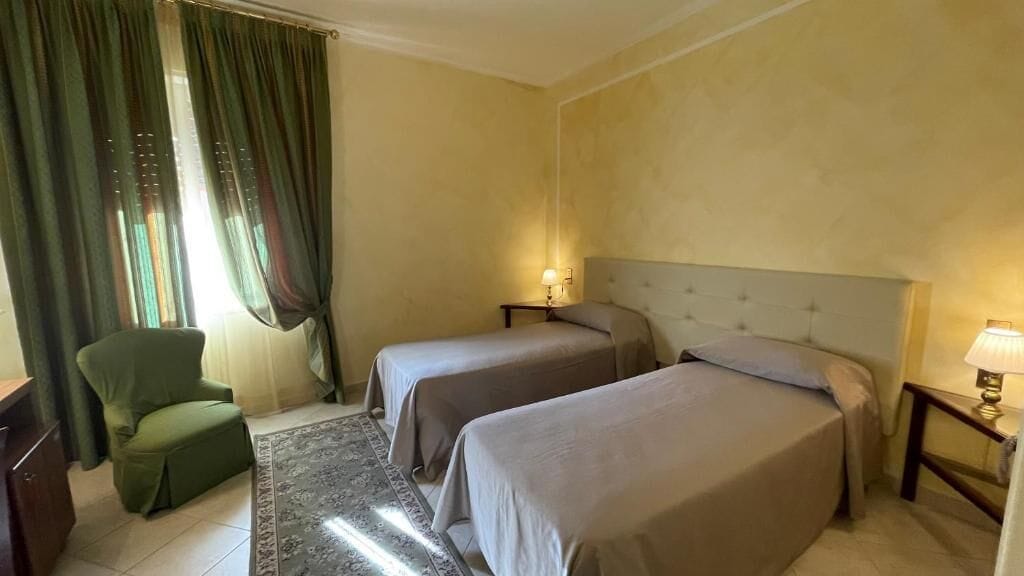 Habitación de hotel con dos camas, un sillón verde, una mesita con lámpara y una ventana con cortinas.