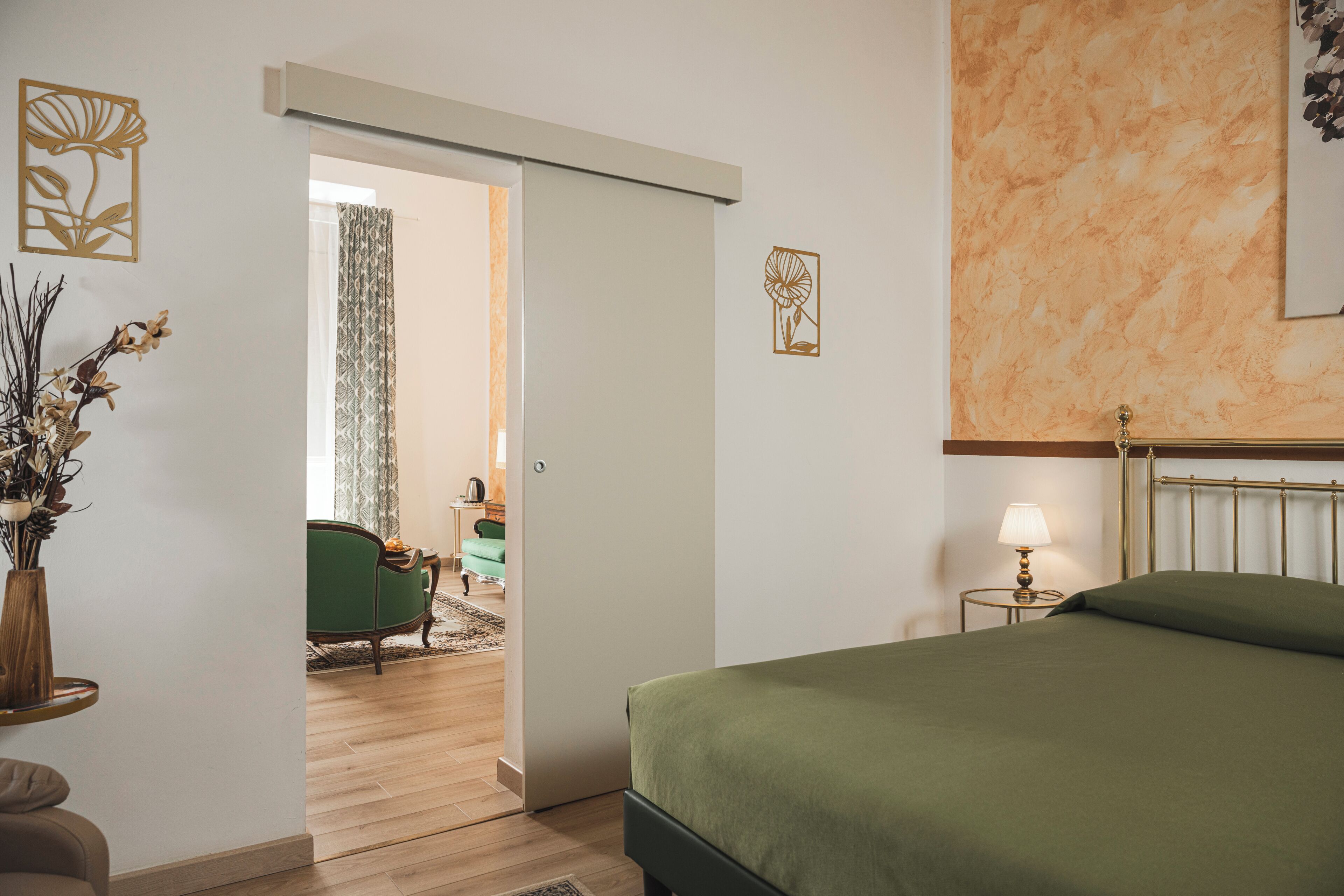 A bedroom with a bed, a nightstand with a lamp, a green chair, a small table with a vase, and a door leading to another room.