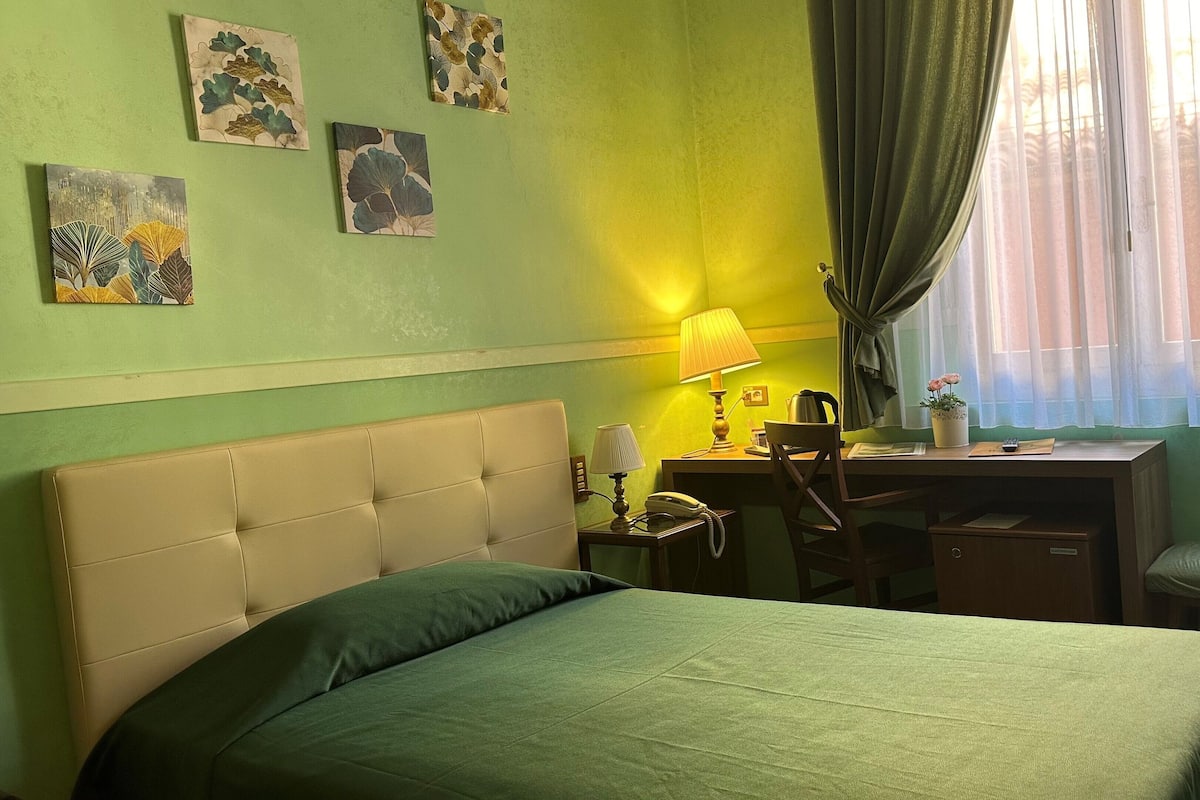 Habitación de hotel con cama, escritorio, silla, lámpara y ventana con cortinas.