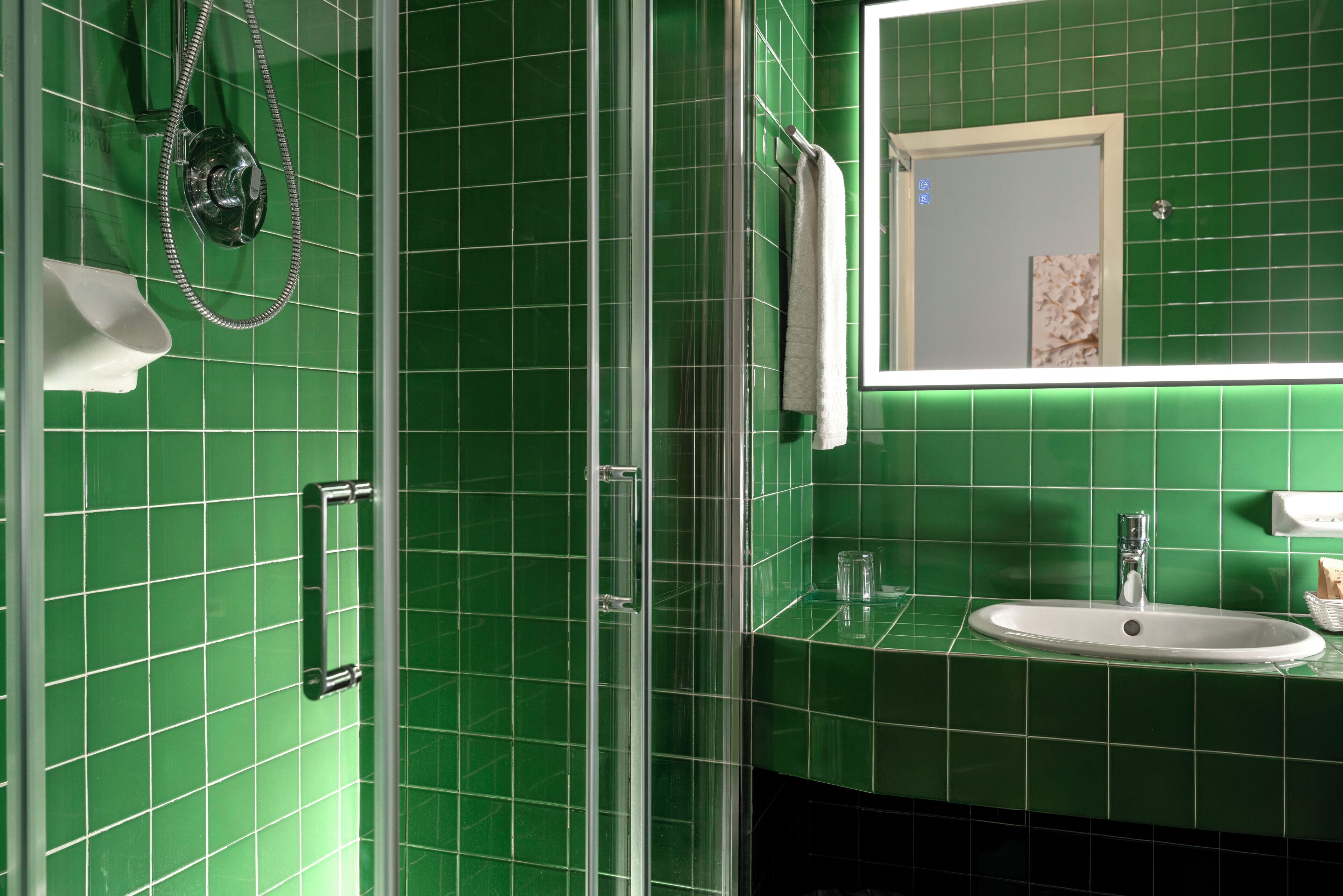 A bathroom with green tiles, a glass shower enclosure, a white sink, and a mirror with a towel hanging on the wall.