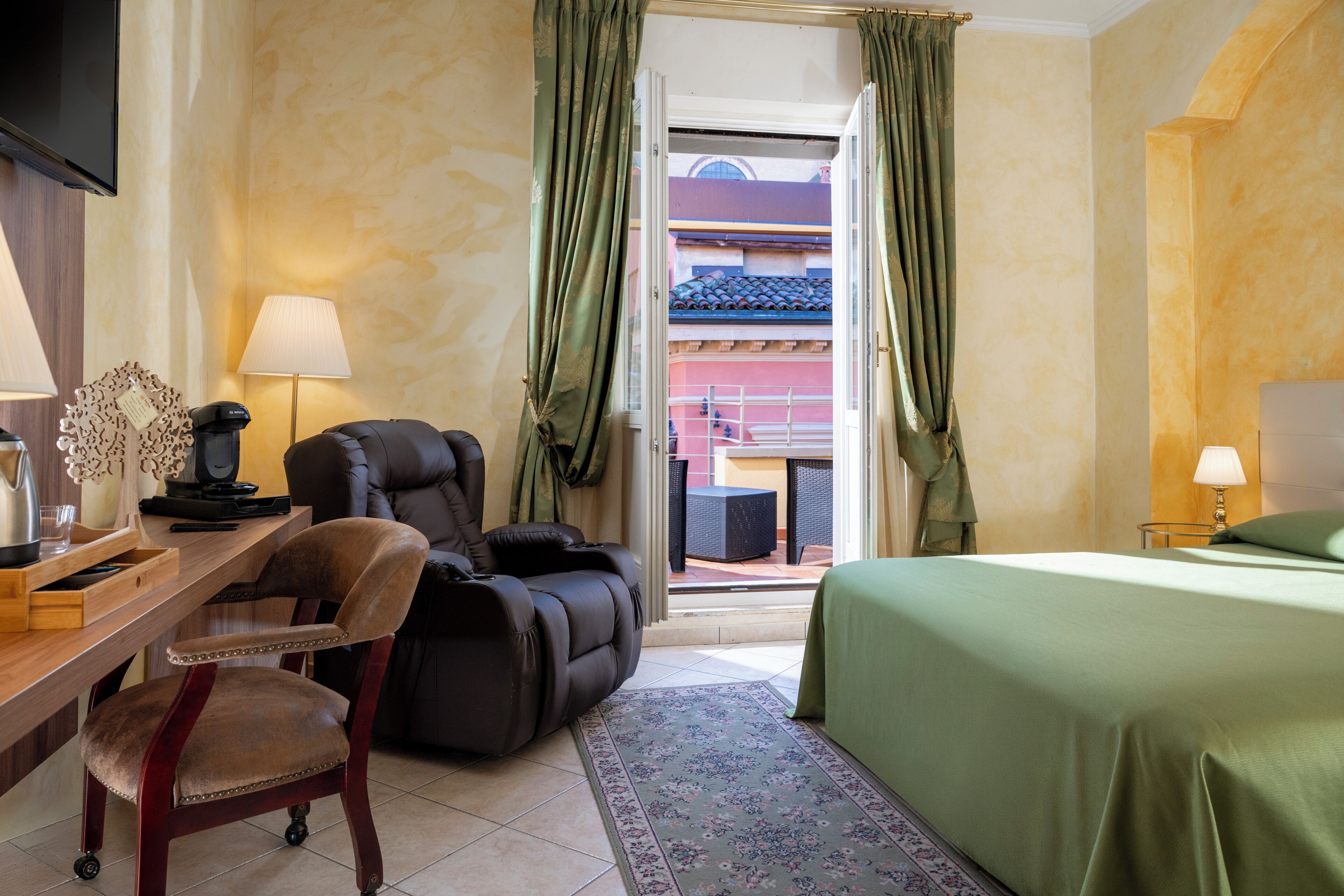 A hotel room with a massage chair, a desk with a lamp, a bed, and a view of a building through the window.