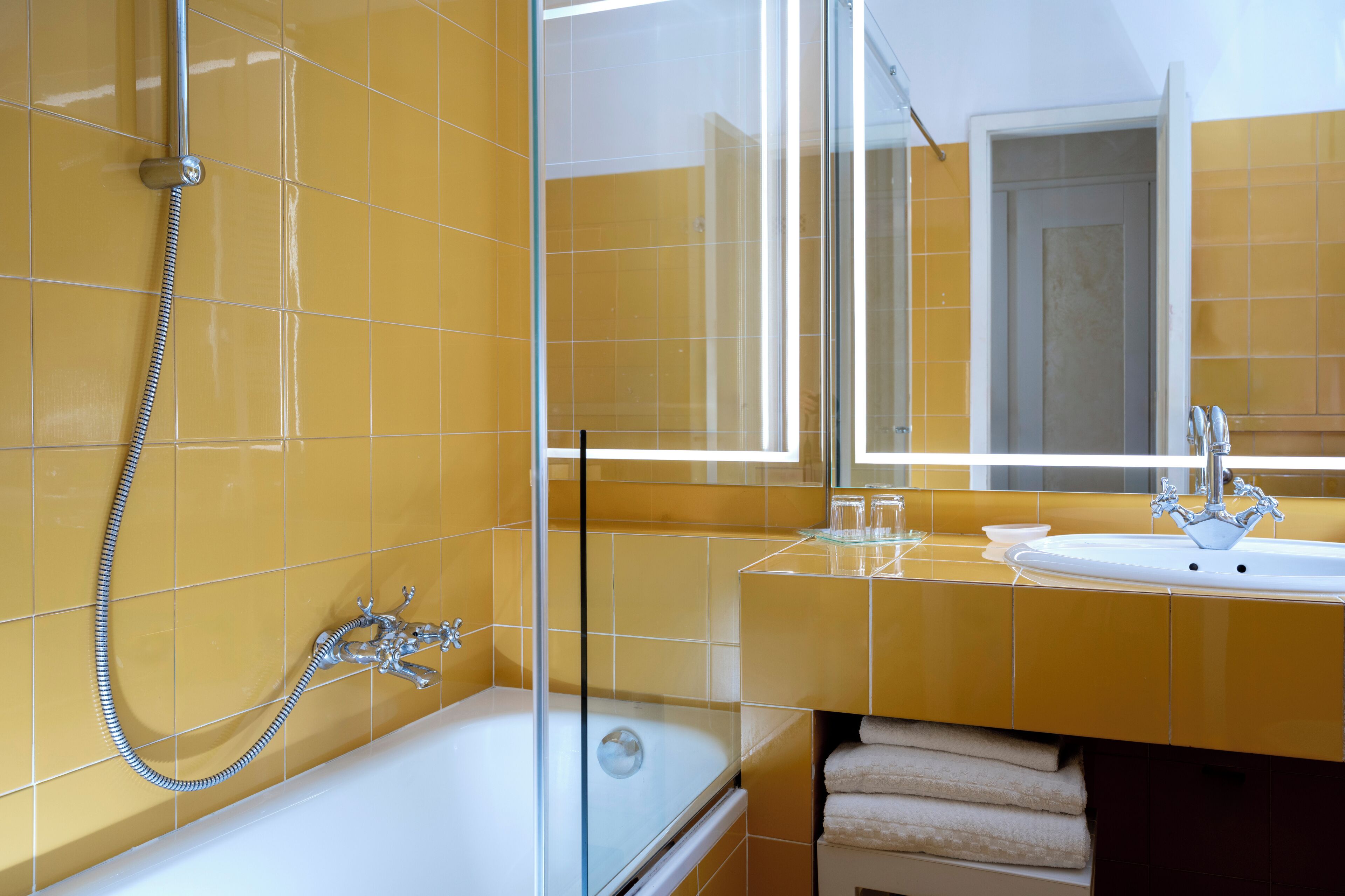Un baño con baldosas amarillas, una ducha con cerramiento de vidrio, una bañera blanca, un lavabo con espejo y toallas ordenadas.