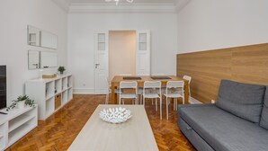 Un salon moderne avec un plancher en bois, un canapé gris, une table à manger avec des chaises blanches et un lustre.