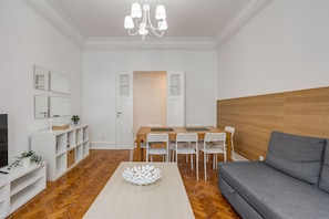 Un salon moderne avec un plancher en bois, un canapé gris, une table à manger avec des chaises blanches et un lustre.