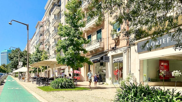 Une rue avec une piste cyclable verte, des terrasses et des devantures de magasins, dont une affiche bien visible avec le mot « CAROL ».