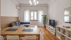 Un salon moderne avec une table à manger en bois, des chaises blanches, un canapé gris, un téléviseur et une bibliothèque.