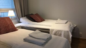 Dos camas individuales con sábanas blancas y almohadas rojas estampadas, una lámpara y una ventana con cortinas.